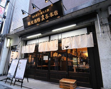 黒と金の看板に白いのれんがかかった「麺屋居酒屋 まる」の外観の写真