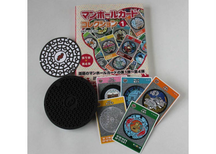 From the right is the book comprised of the 222 Manhole Cards, and next are five manhole cards. To the left is an iron coaster based on a 1/6 manhole replica bearing a type of hexagonal pattern called Bishamon kikkō pattern, and a paper coaster featuring the standard JIS pattern which enthusiasts call a “plain cover”.