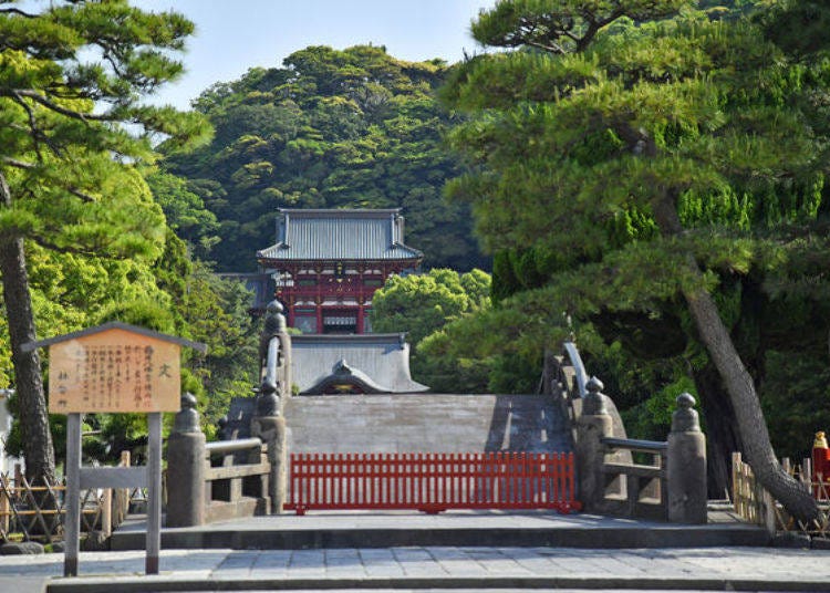 A shrine is nestled in deep greenery behind the arched bridge.