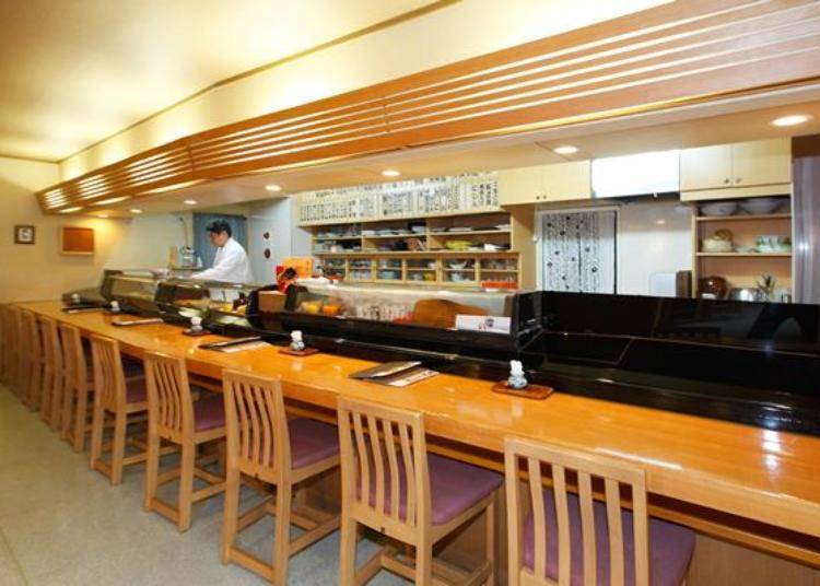 There are also tables and tatami mat seating in addition to the counter. Manager Masaki Yamamoto is pictured standing in the kitchen area.