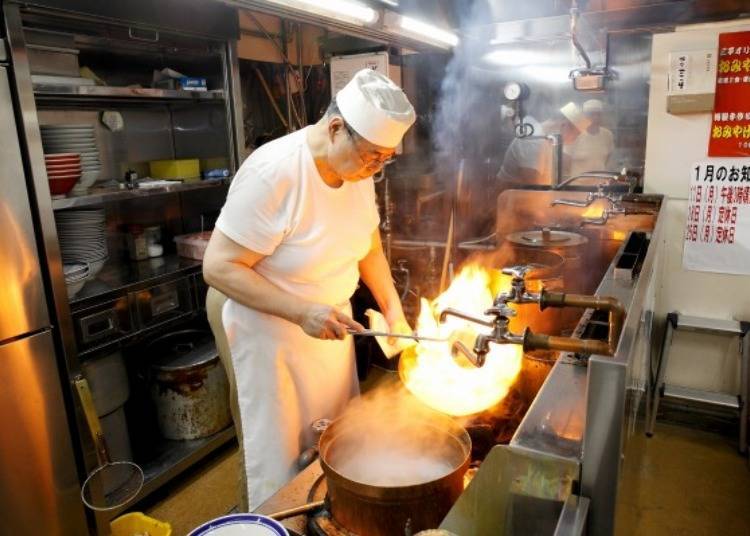 ▲ While steaming noodles, he stir-fries plenty of vegetables in a wok. The chef depicted here is third generation descendant, Kazuto Omiya.