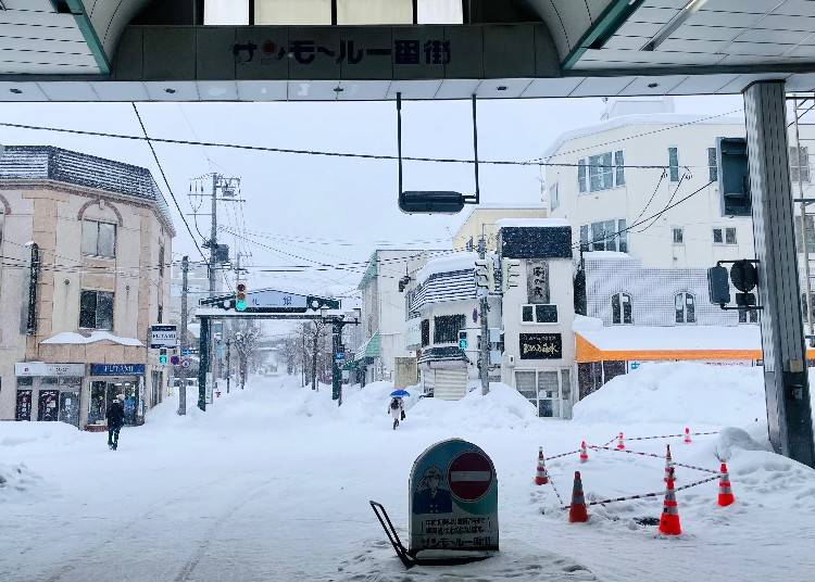▲澤の露本舗は小樽駅から徒歩約10分、サンモール商店街（写真手前側）を抜け信号を渡った先の細長い建物