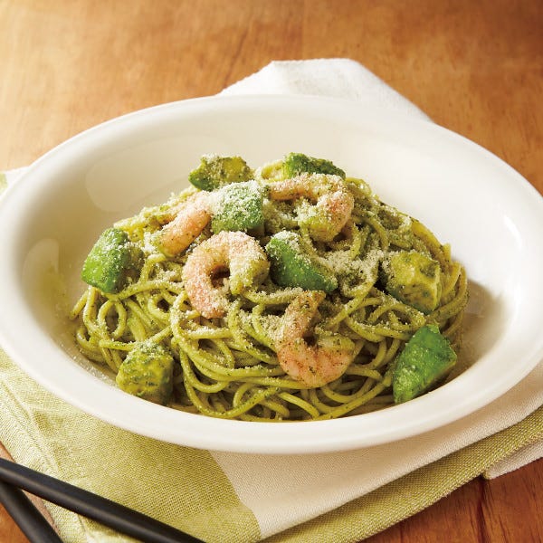 木製のテーブルの上に、白い丸皿に盛り付けられた海老とアボカドのバジルソース生パスタが置かれている