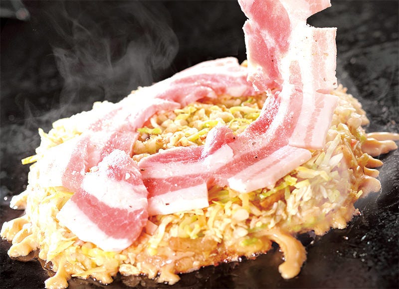 鉄板の上で焼かれているお好み焼き。たっぷりのキャベツの上に新鮮な豚肉がのせられている。